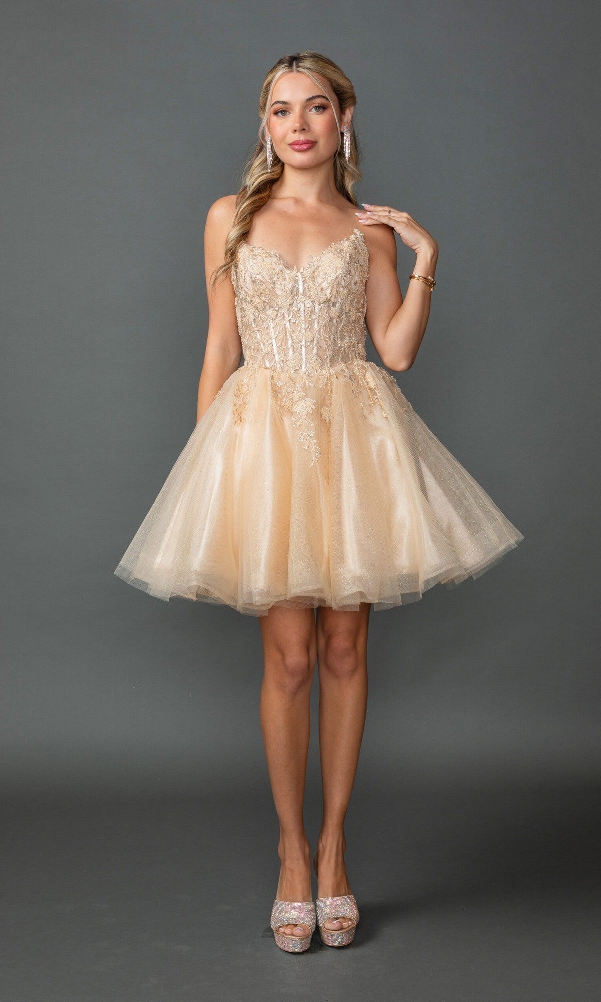A woman stands against a gray background wearing the Dancing Queen 3431 short homecoming dress in champagne, featuring a cream-colored lace embroidered corset bodice and an elegant tulle A-line skirt, paired with high heels.